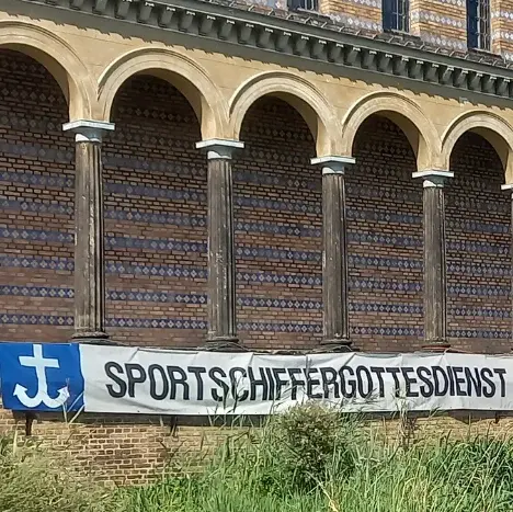 Ein Gebäude direkt am Wasser mit vielen Säulen und einem Banner mit der Aufschrift ,,Sportschiffergottesdienst''.