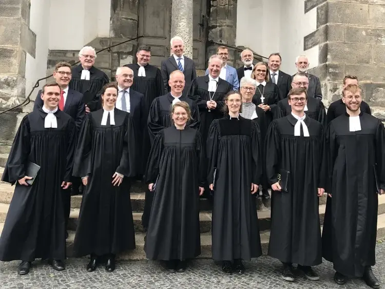 Alle Beteiligten des Ordinationsgottesdienstes 2022 in Talar und Anzügen. Sie stehen auf einer Kirchentreppe in Görlitz und lächeln in die Kamera.