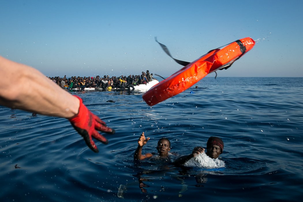 Zwei Geflüchtete, die nach einem Bootsunglück im Wasser schwimmen und einen Rettungsring zugeworfen bekommen. Im Hintergrund ein überfülltes Schlauchboot.