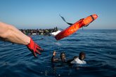 Zwei Geflüchtete, die nach einem Bootsunglück im Wasser schwimmen und einen Rettungsring zugeworfen bekommen. Im Hintergrund ein überfülltes Schlauchboot.