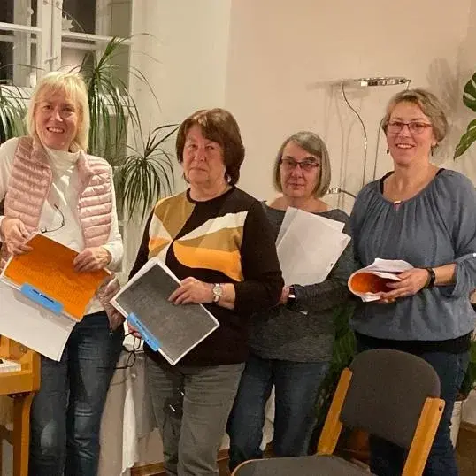 Vier Frauen stehen in einem Raum neben einem Klavier und halten beschriftete Blätter in der Hand.