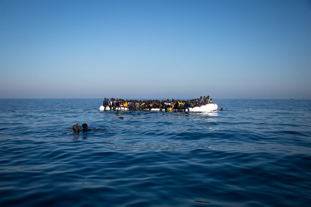 Geflüchtete Menschen, die neben einem überfüllten Schlauchboot im Wasser im offenen Meer schwimmen.