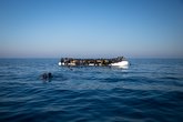Geflüchtete Menschen, die neben einem überfüllten Schlauchboot im Wasser im offenen Meer schwimmen.