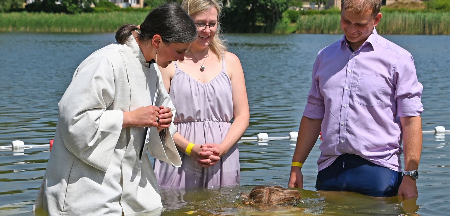 Ein Kind taucht bei seiner Taufe in einem See unter. Daneben stehen die Eltern und die Pfarrerin im weißen Gewand.