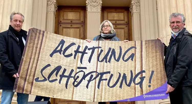Bischof Stäblein, Amet Bick und Hans Georg Baaske halten ein Banner mit der Aufschrift ,,Achtung Schöpfung''.