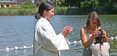 Ein Mädchen, das gerade in einem See getauft wurde. Die Mutter hält lächelnd ihren Kopf und daneben steht die Pfarrerin und klatscht.