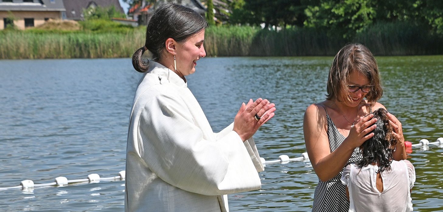 Ein Mädchen, das gerade in einem See getauft wurde. Die Mutter hält lächelnd ihren Kopf und daneben steht die Pfarrerin und klatscht.