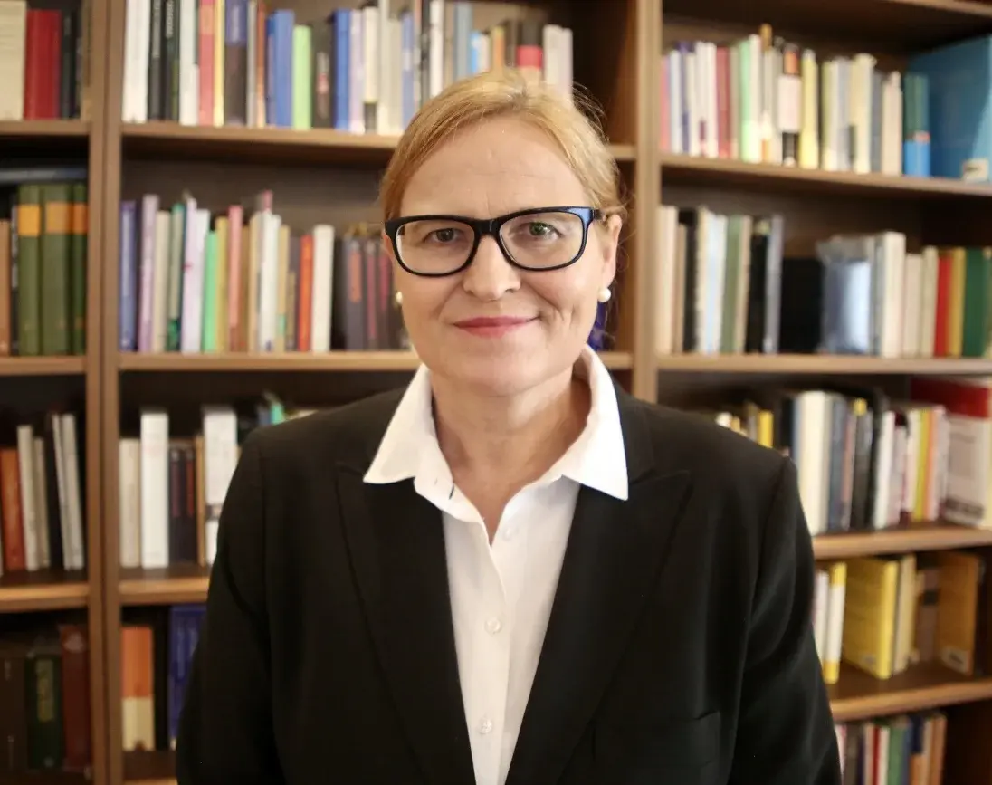 Ulrike E. Auga steht vor einem Bücherregal und schaut in die Kamera.