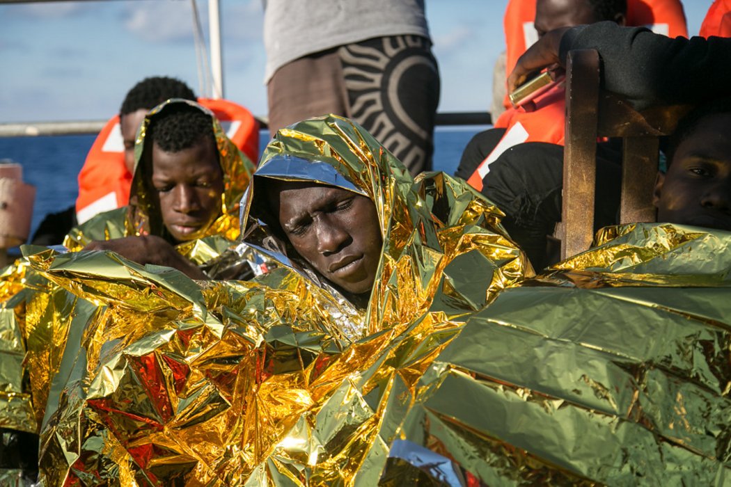 Geflüchtete Menschen, die nach einem Bootsunglück mit Rettungsdecken und Schwimmwesten auf einem Rettungsboot sitzen und die Augen schließen.