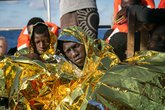 Geflüchtete Menschen, die nach einem Bootsunglück mit Rettungsdecken und Schwimmwesten auf einem Rettungsboot sitzen und die Augen schließen.