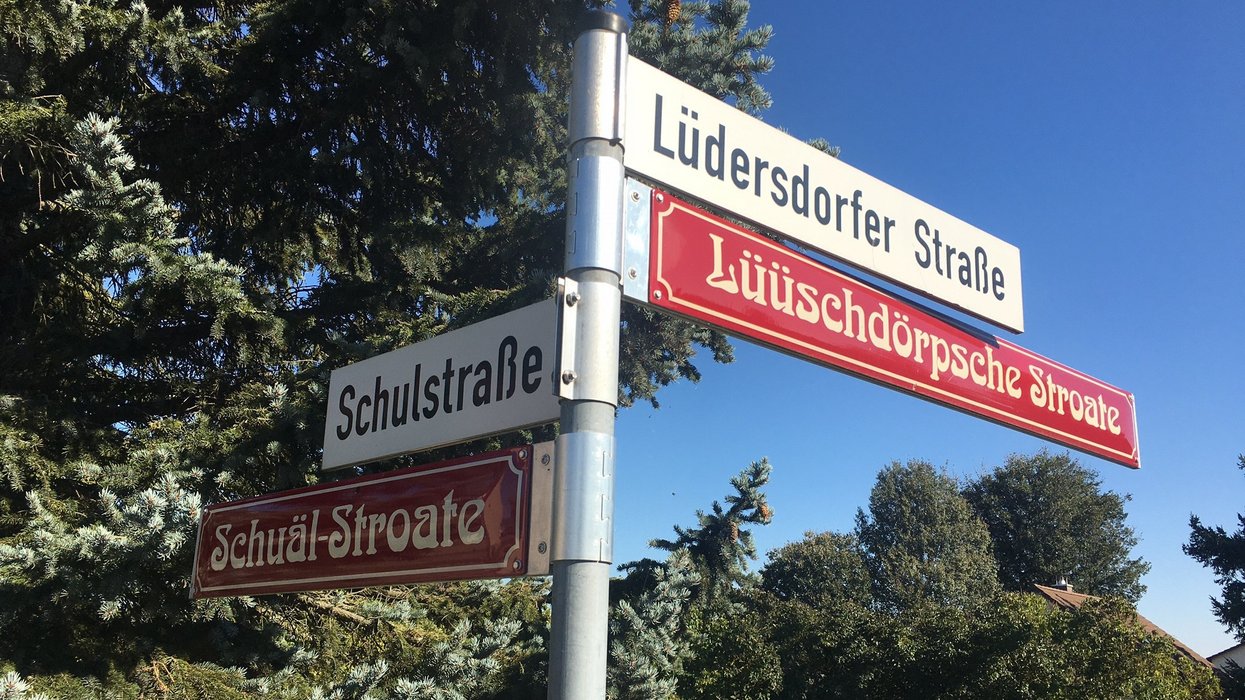 Straßenschilder von zwei Straßen, die jeweils mit einem hochdeutschen und einem plattdeutschen gekennzeichnet sind. Im Hintergrund Bäume und blauer Himmel.