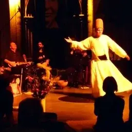 Bühnenbild, auf dem sich ein Mann in einem hellen Gewand bewegt. Im Hintergrund sitzen Personen mit Instrumenten.