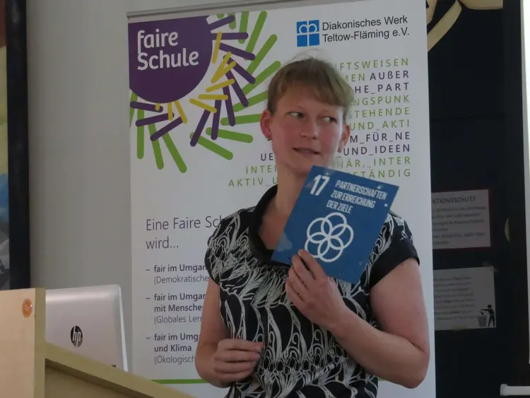 Eine Frau steht vor einem ,,Faire Schule'' Werbebanner und hält eine blaue Karte mit der Nummer 17 und der Beschriftung ,,Partnerschaften zur Erreichung der Ziele'' in der Hand.
