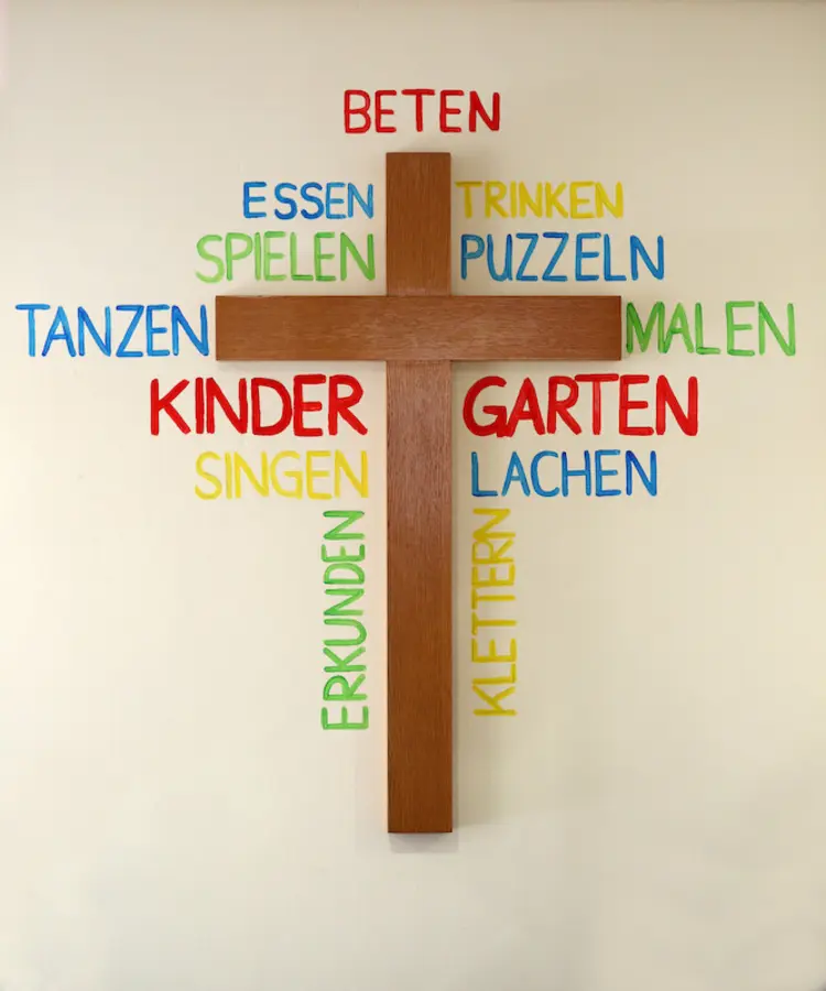 Holzkreuz, das an einer Wand hängt und von bunten Wörtern umgeben ist.