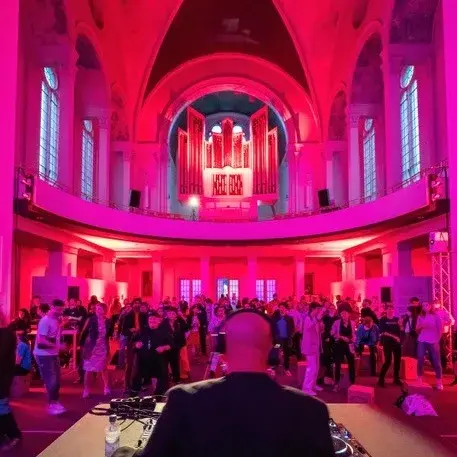 Eine Gruppe von Menschen tanzt in einer rosa beleuchteten Kirche bei dem Electronic Church Event. Im Vordergrund steht ein DJ an seinem Pult.