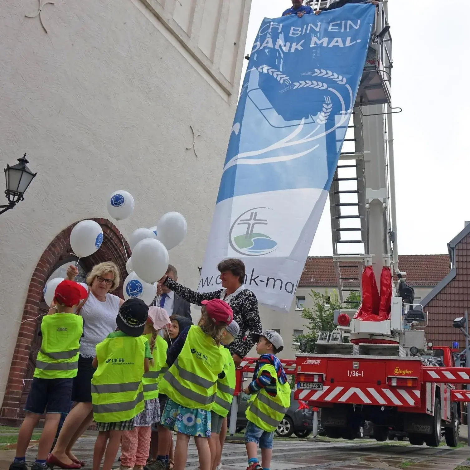 Eine Gruppe von Kindern und drei Erwachsenen steht mit Luftballons in der Hand vor einem Gebäude. Im Hintergrund steht eine Feuerwehr, an der ein Banner mit der Aufschrift ,,Dank mal'' hängt.