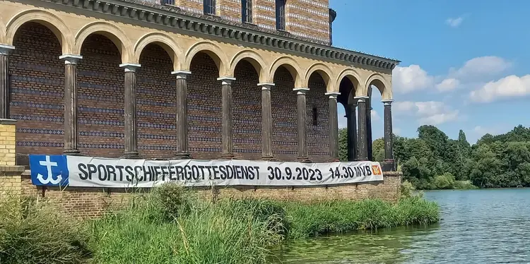 Ein Gebäude direkt am Wasser mit vielen Säulen und einem Banner mit der Aufschrift ,,Sportschiffergottesdienst''.