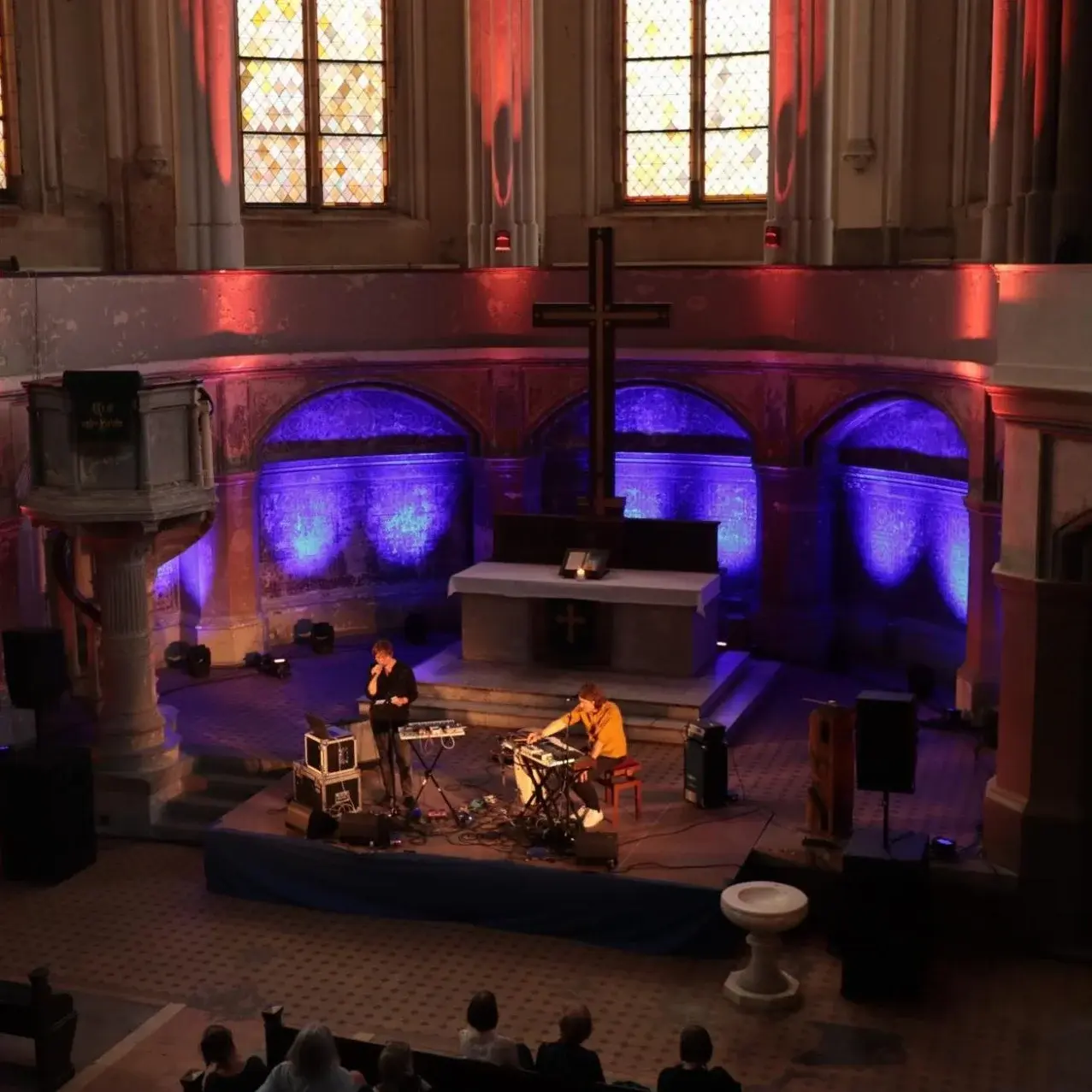 Rot und violett beleuchtete Kirche mit Publikum. Vorne sind zwei Personen mit Keybords und Mikrofon zu sehen.