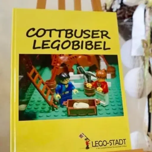 Gelbes Cover der Cottbuser Legobibel, das ein Legobild mit zwei Figuren und verschiedenen Gegenständen zeigt.