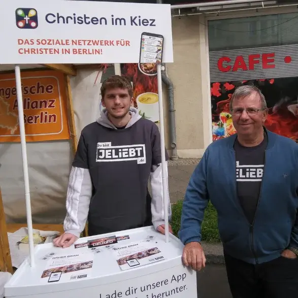 Zwei Männer stehen neben und hinter einem Stand, auf dem ,,Christen im Kiez'' steht. Sie tragen T-shirts mit der Aufschrift ,,Jeliebt''.