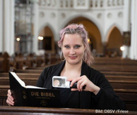 Eine Frau Mitte 20 sitzt in einer Kirchenbank und lächelt in die Kamera, in einer Hand eine Leuchtlupe, in der anderen eine Bibel. Sie hat langes, dunkelblondes Haar mit rötlichen Strähnchen und trägt große Ohrringe.