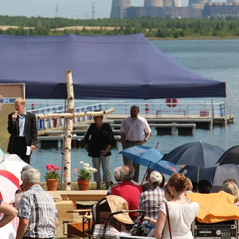 Mehrere Personen sitzen mit Bickrichtung zu einem See. Vor ihnen stehen drei Personen, einer von ihnen spricht in ein Mikrofon. Neben ihm steht ein großes Kreuz aus Baumstämmen.