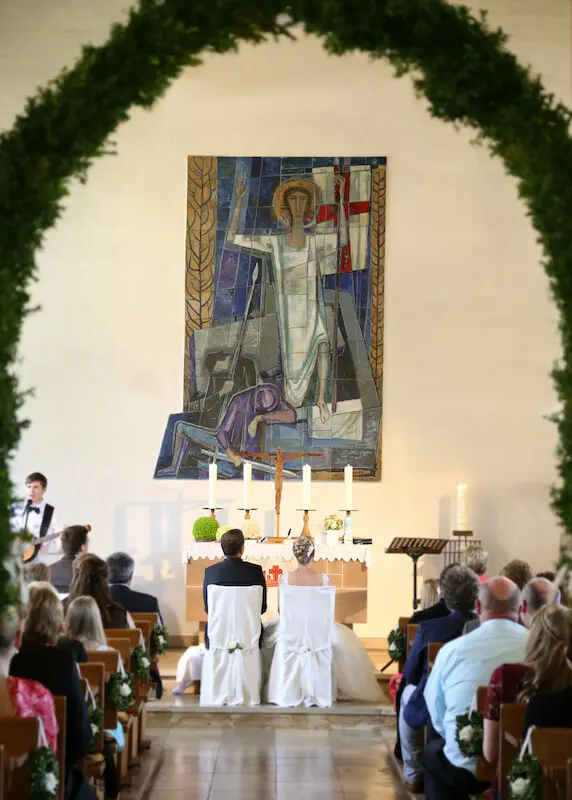 Ehepaar sitzt auf Stühlen vor dem Altarraum. Braut in weißem Kleid. Bräutigam links daneben in schwarzem Anzug. Links sieht man die Band.