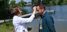 Ein Mann erhält den Taufsegen von einer Frau am Ufer eines Sees. Im Hintergrund ist ein Steg und eine Frau mit Kamera.