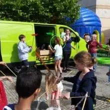 Mehrere Kinder spielen mit verschiedenen Gegenständen auf einem Gehweg. Im Hintergrund steht ein hellgrüner Kleinbus, eine Hüpfburg und zwei Erwachsene.
