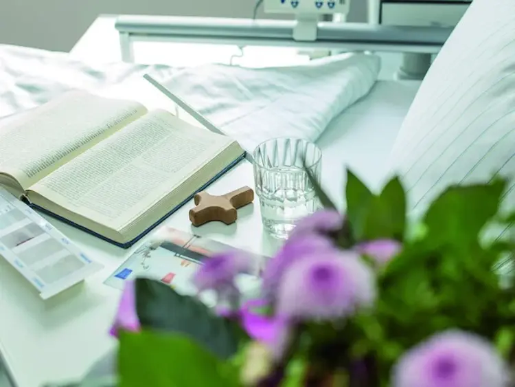 Krankenbett, an dem ein kleines Holzkreuz, eine Bibel, Medikamente, Wasser und Blumen stehen.