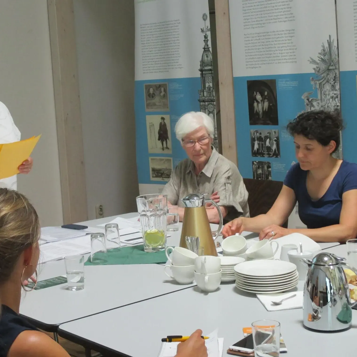 Drei Frauen sitzen an einem mit Geschirr und Kannen gedeckten Tisch. Vor ihnen liegen Stift und Papier. Am Ende des Tisches steht eine Frau mit einem gelben Zettel in der Hand.