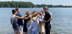 Eine Gruppe von Personen steht in einem See. Eine von ihnen hält ein Baby auf dem Arm und die anderen legen ihre Hand auf den Kopf des Kindes. 
