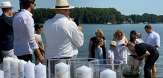 Mehrere Personen stehen in einem See. Ein Pfarrer hält ein wenig Wasser in einer Hand, in der anderen ein Buch. Darunter, auf dem Wasser, liegt ein Blumenkranz.