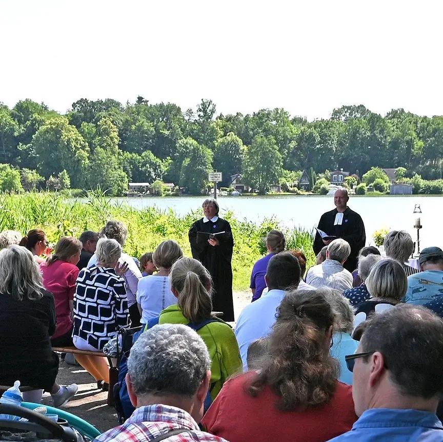 Eine Gruppe von Menschen sitzt an einem See auf Bänken in Blickrichtung zu zwei Pfarrern, die zu den Menschen sprechen.