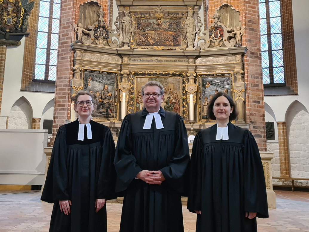 Drei Menschen stehen nebeneinander vor einem Altar in einer Kirche. Alle tragen einen Talar