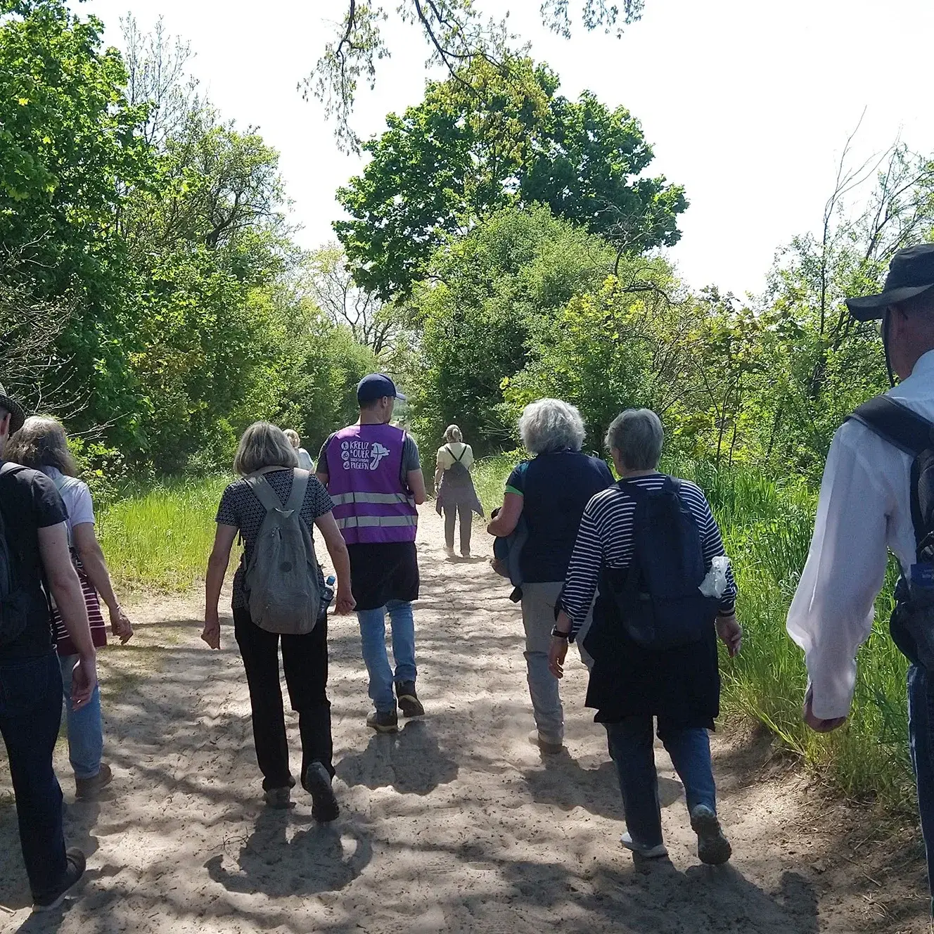 Eine Gruppe von Menschen geht den Spandauer Pilgerweg entlang, der umgeben ist von grünen Bäumen und Büschen. 