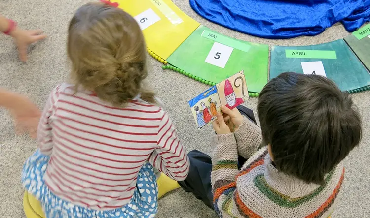 Kinder sitzen auf dem Boden. Eins von ihnen hält Karten mit Bildern in der Hand. Vor ihnen liegt ein bunter Kreis, auf dem die Monate des Jahres stehen.