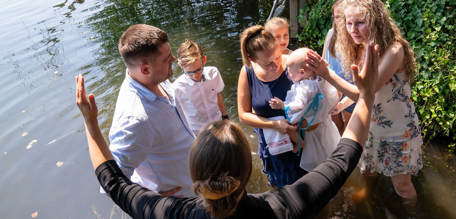 Ein Baby sitzt auf dem Arm einer Frau und wird getauft. Daneben stehen mehrere Personen und eine schwarz gekleidete Frau, die ihre Hände empfangend in die Luft hält.