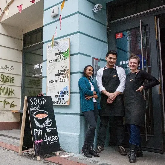Drei Personen stehen vor dem Eingang eines Gebäudes. Zwei von ihnen tragen eine schwarze Schürze. An der Hauswand neben ihnen steht ein Schild, das Suppe für 4 Euro anbietet.