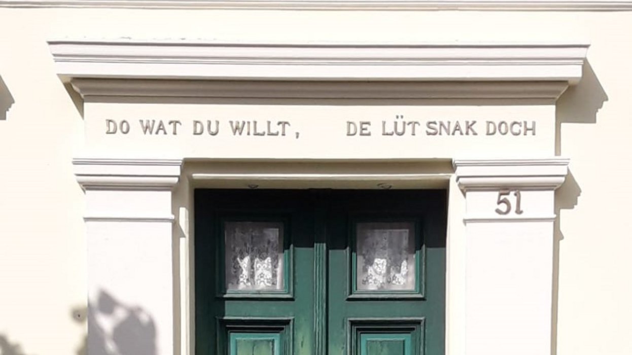 Hauswand mit grüner Eingangstür, über der ,,do wat du willt, de lüt snak doch'' geschrieben steht.
