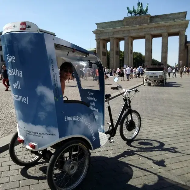 Eine Person sitzt in einer Rikscha und schaut zur Kamera. Im Hintergrund ist das Brandenburger Tor und blauer Himmel zu sehen.