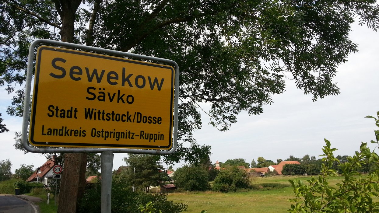 Ortseingangsschild des Ortes Sävko, das am Straßenrand steht. Im Hintergrund eine Wiese, Bäume, Sträucher und Häuser.