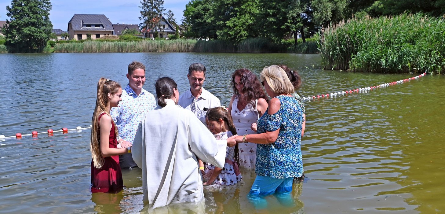 Sieben Personen stehen in einem Kreis im See und halten sich an den Händen. In ihrer Mitte stehen Kinder.
