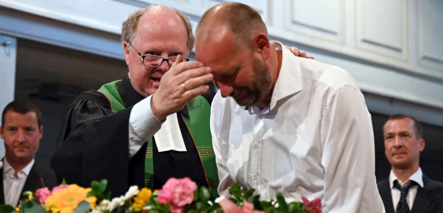 Ein Mann in einem weißen Hemd wird vom Pfarrer getauft. Im Hintergrund stehen zwei Männer im Anzug.