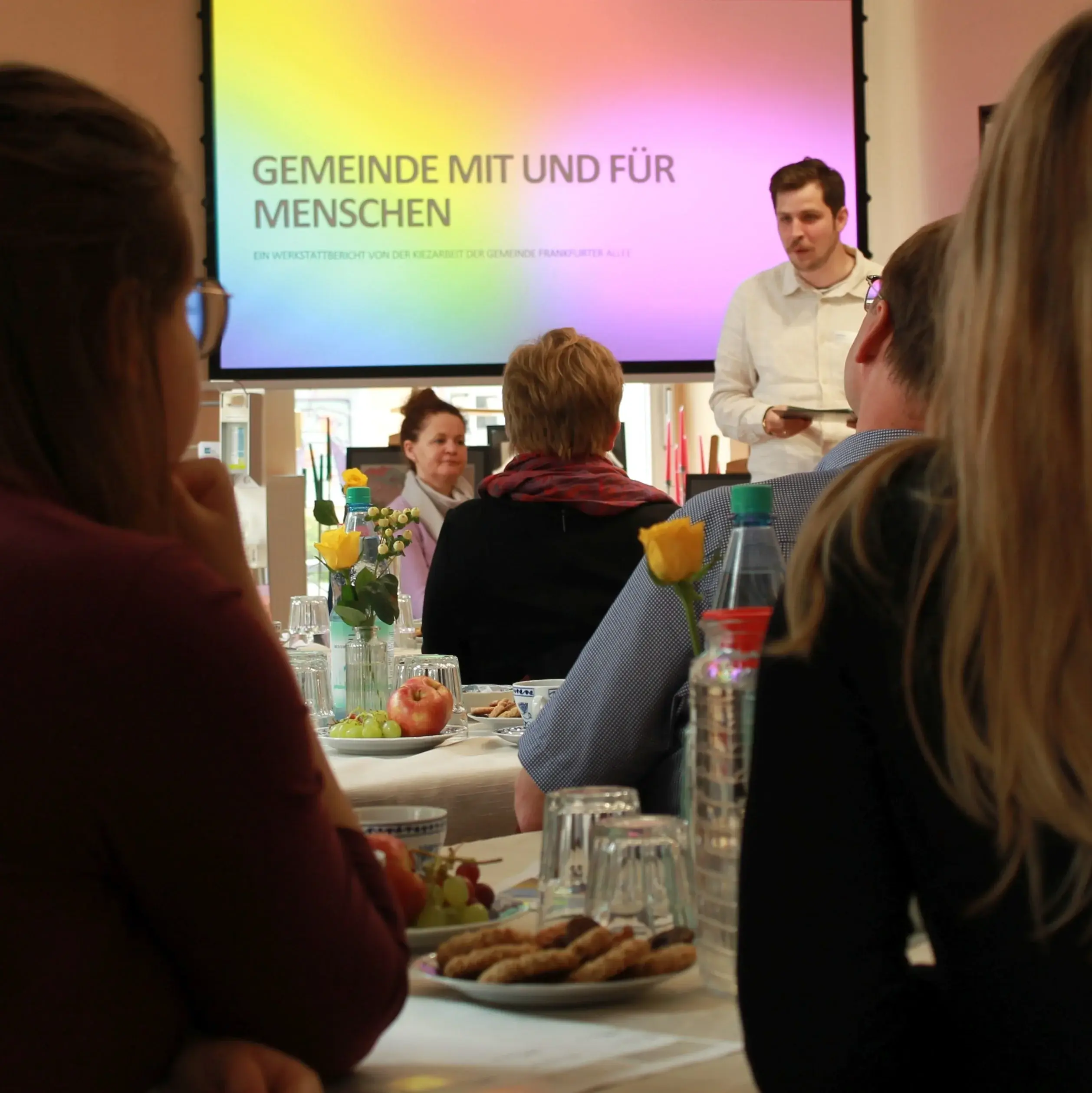 Ein Mann hält eine Präsentation im Kiezcafe ,,ein Laden''. Mehrere Menschen sitzen an Tischen mit Gebäcken und Wasser und hören zu.