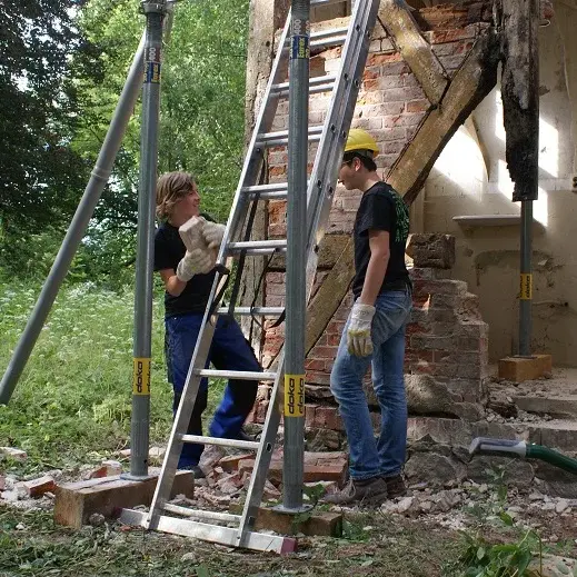 Zwei Jugendliche stehen vor einem zerfallenen Haus. Einer von ihnen hält einen Ziegelstein. Um sie herum ist ein Baustellengerüst gebaut.