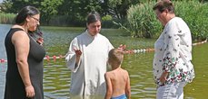 Ein Junge wird von einer Frau im weißen Gewand in einem See getauft. Daneben stehen zwei weitere Frauen und schauen zu.