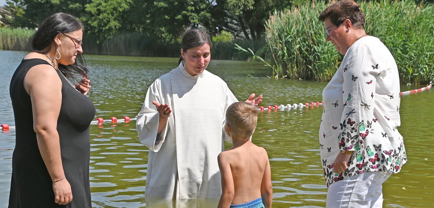 Ein Junge wird von einer Frau im weißen Gewand in einem See getauft. Daneben stehen zwei weitere Frauen und schauen zu.