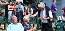 Eine ältere Dame sitzt an einem See auf einem Stuhl und erhält einen Taufsegen. Im Hintergrund stehen weitere Personen.