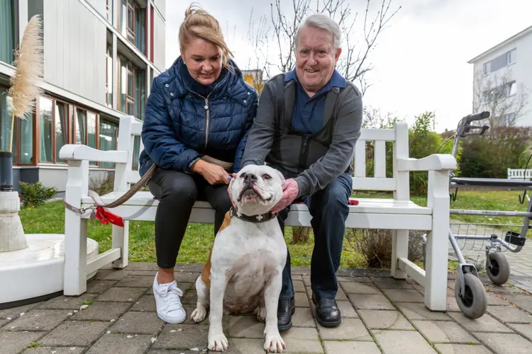 Zwei Senioren sitzen auf einer Bank und streicheln einen Hund.